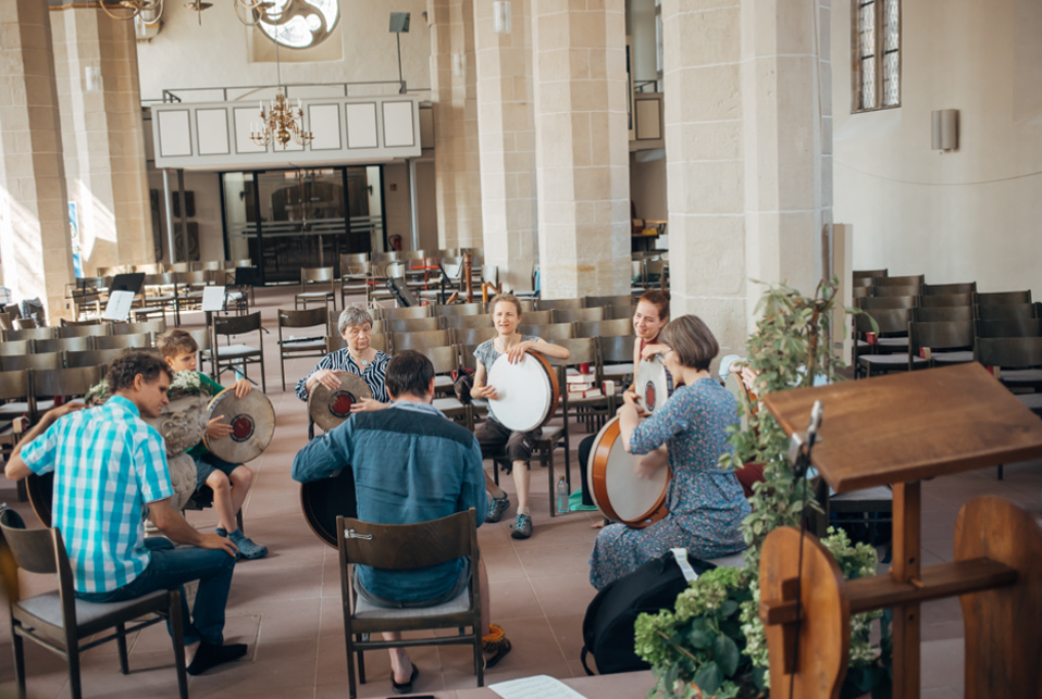 Eine Gruppe junger und älterer Menschen hat in einem Kirchenraum einen Kreis gebildet. Sie trommeln gemeinsam.
